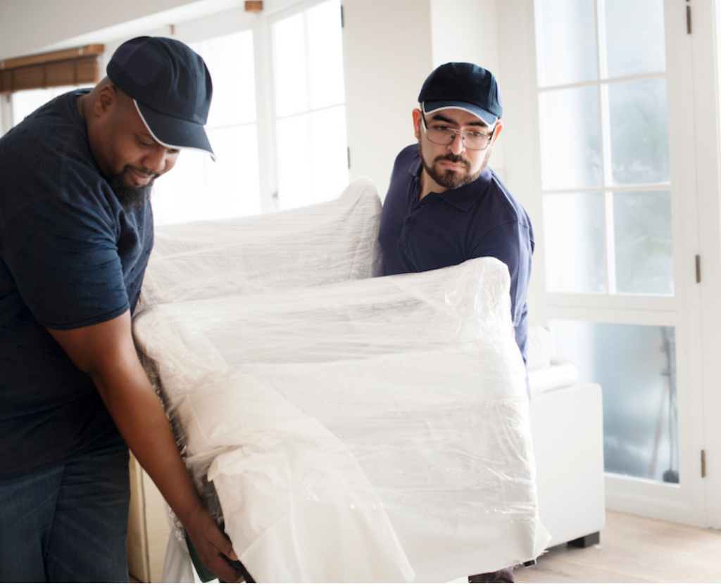 Two delivery workers carefully moving a wrapped piece of furniture into a bright, modern room—illustrating professional FF&E delivery and installation service.
