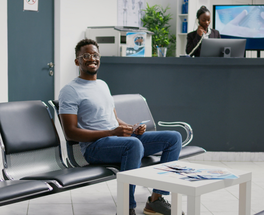 Young male patient smiling while seated in a modern clinic lobby with metal-framed chairs, a white coffee table, and a receptionist working in the background.