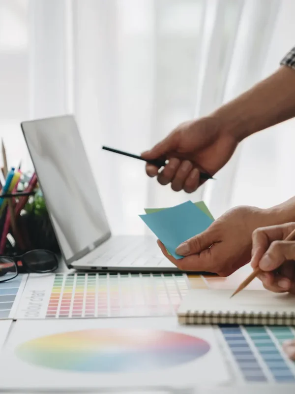 Designers reviewing color swatches and palette samples at a desk with a laptop, notebook, and color charts—planning a layout for an interior FF&E project.